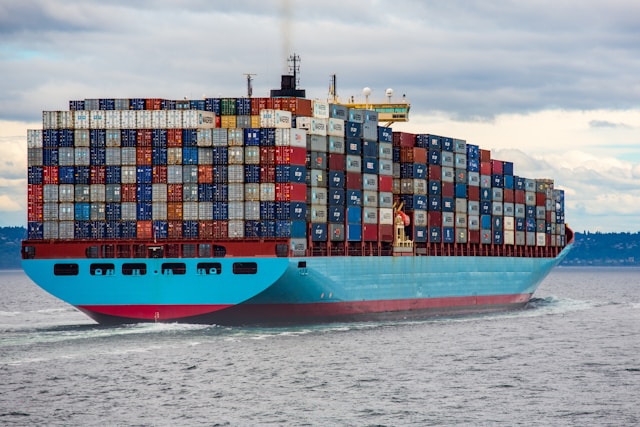 A large cargo ship carrying stacks of multicolored shipping containers travels across a body of water under a cloudy sky.